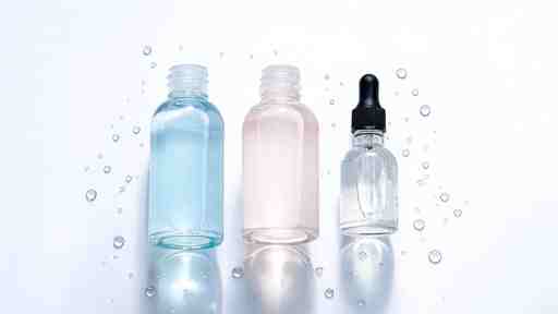 Flat lay of glass-skin Korean skincare — hydrating toner, essence, and hyaluronic acid serum — on a glossy reflective surface with water droplets