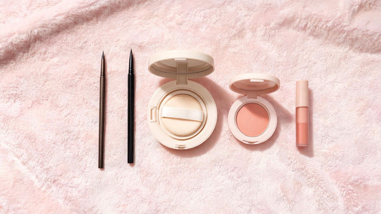 Douyin-style makeup product flat lay with fine brow pencil, thin eyeliner, pale cushion foundation, peachy blush and lip tint on a soft pink surface